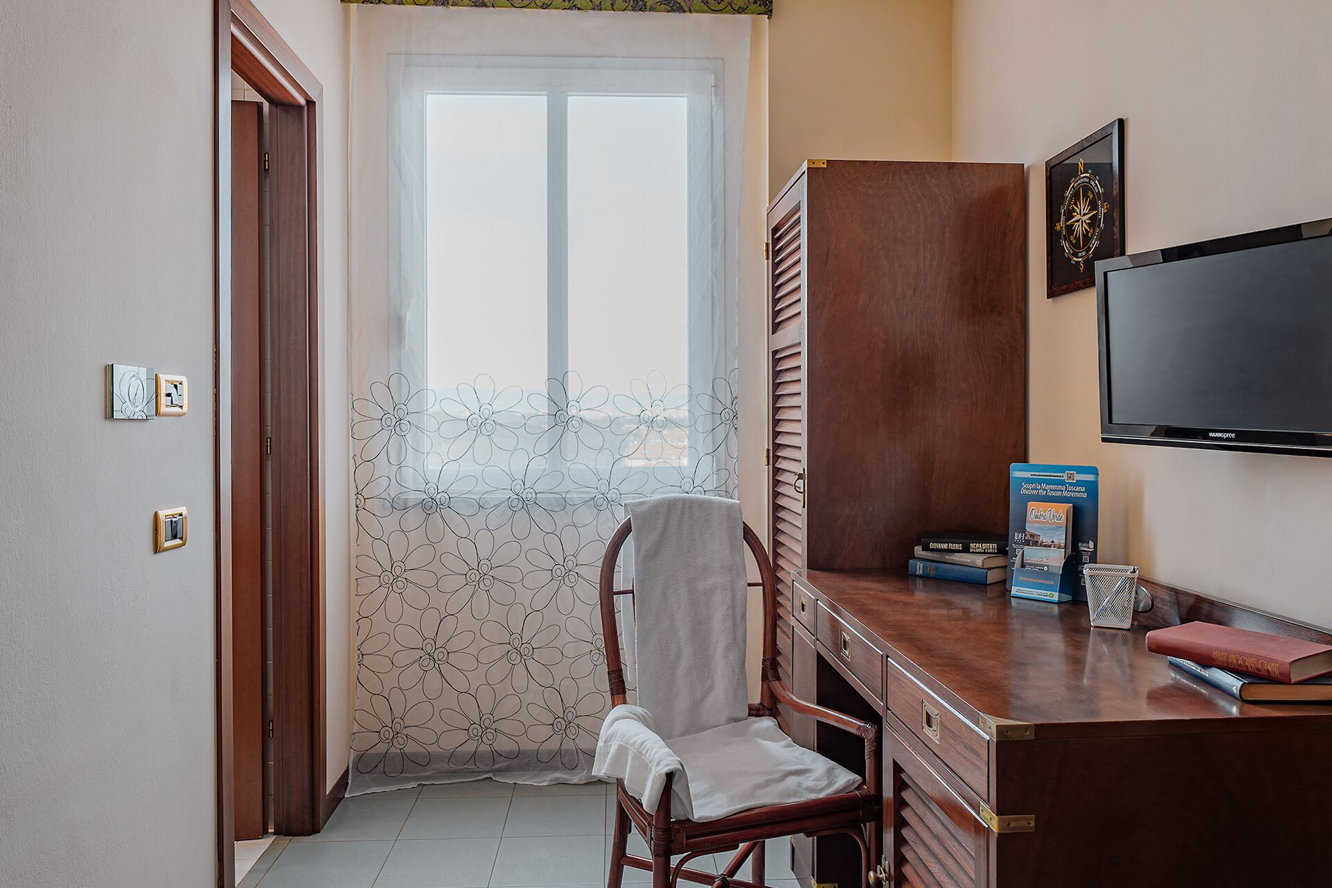 A small room with a wooden desk, a chair, a television, and a window with a floral curtain.