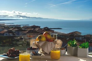 Ein Tisch mit Frühstücksangeboten, ein Glas Orangensaft und Topfpflanzen auf einem Balkon mit Blick auf eine Küstenstadt.