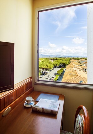 Una habitación con una mesa de madera, una taza y un periódico. Una ventana ofrece vistas a una calle con coches aparcados y árboles.