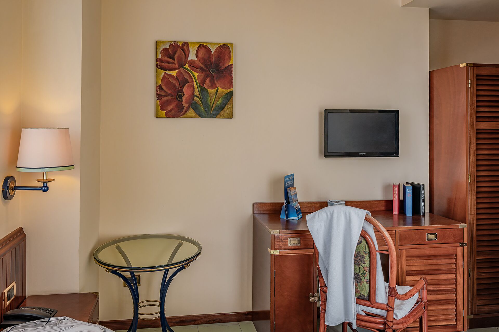 A hotel room with a bed, a desk with a chair, a small table, a wall-mounted TV, and a painting of flowers.