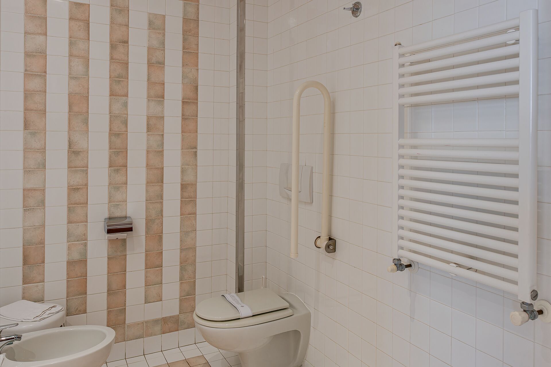Un baño con inodoro, bidé y ducha, con pared de azulejos blancos y un perchero para toallas.