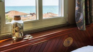 Ein Zimmer mit einem hölzernen Kopfteil, einem Bett mit weißer Bettwäsche, einem Fenster mit Meerblick und einem Glas mit Muscheln auf dem Fensterbrett.