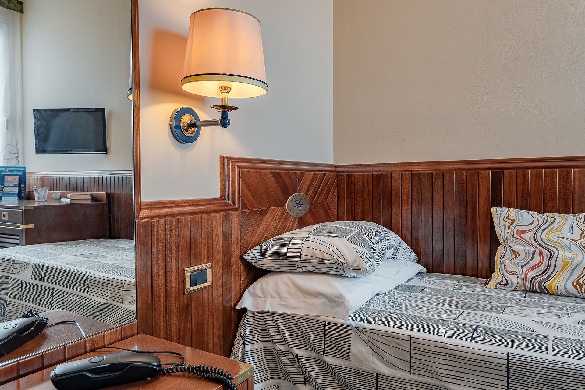 Ein Hotelzimmer mit zwei Betten, einem Nachttisch mit Telefon, einer Lampe und einem an der Wand befestigten Fernseher.