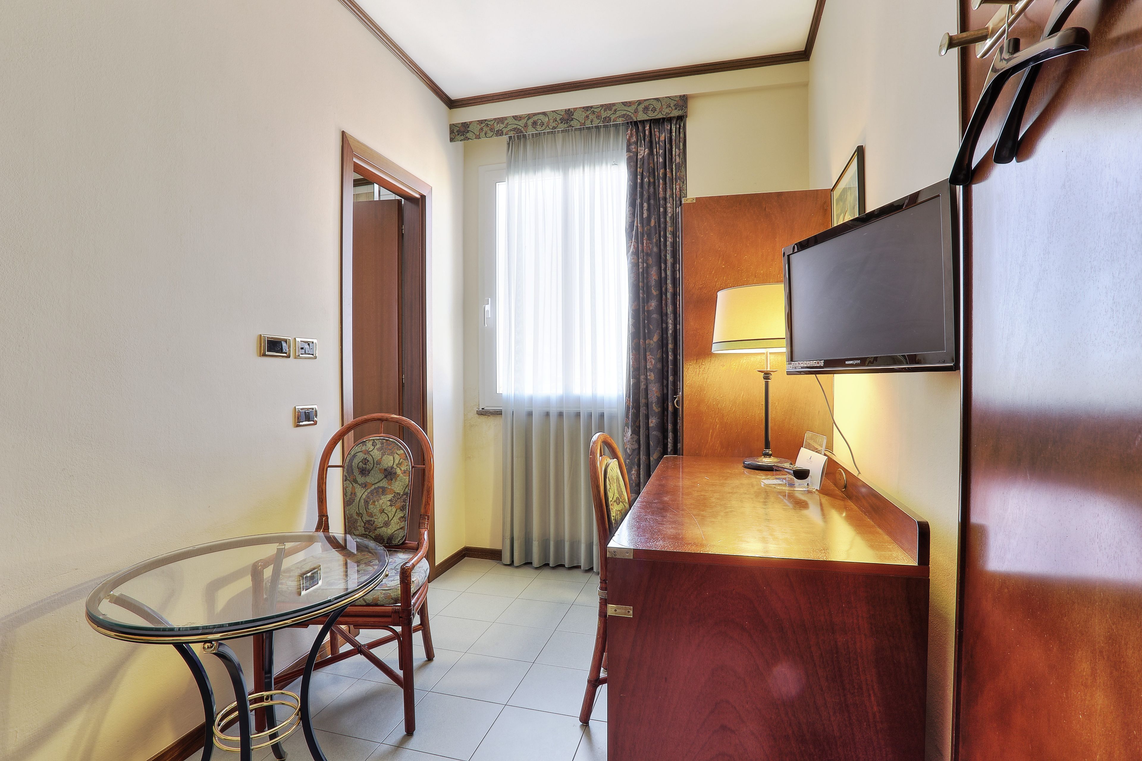 A hotel room with a glass table, two wooden chairs, a wooden desk, a television, and a window with curtains.