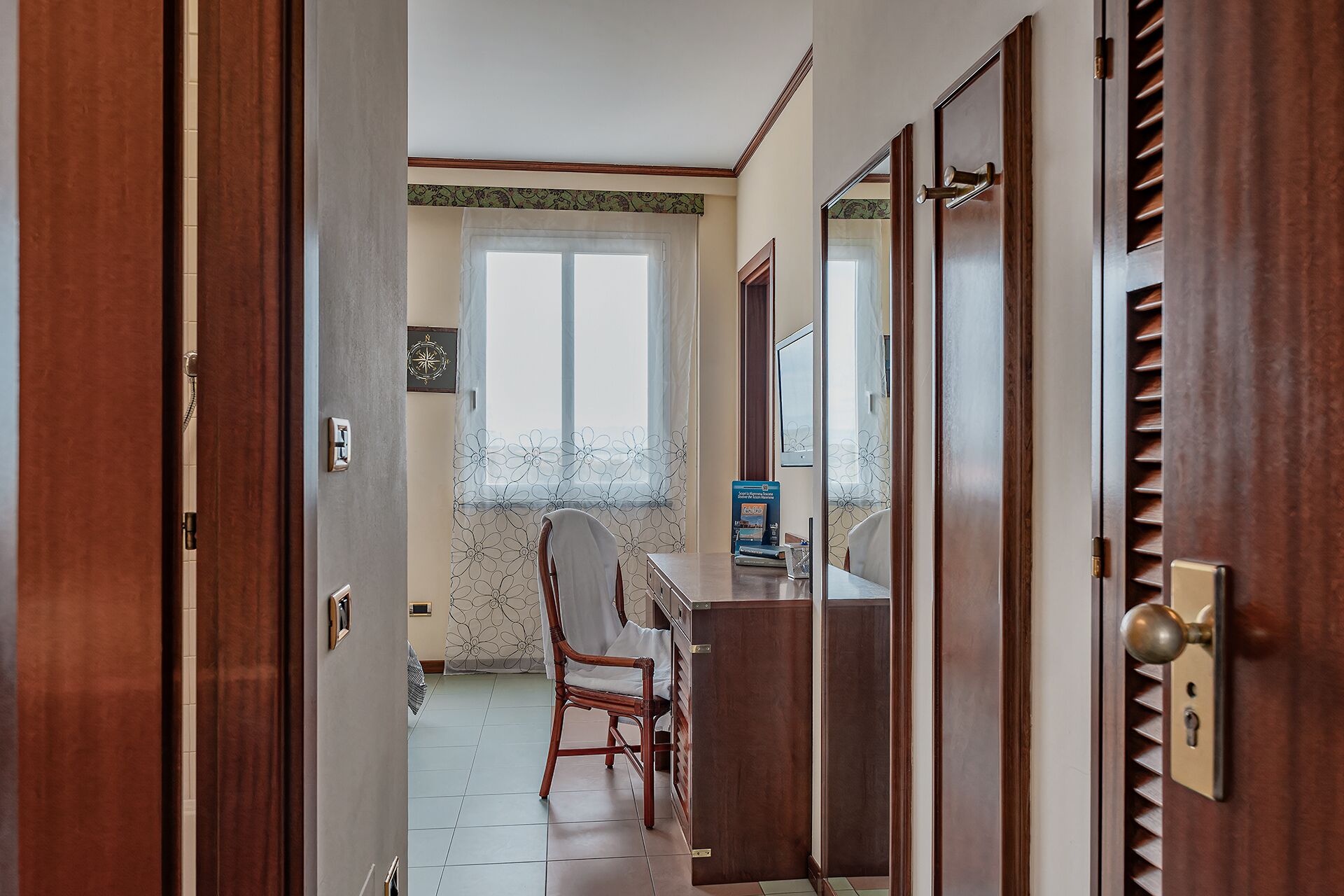 Ein Flur mit einer Holztür, einem Fenster mit blumigem Vorhang, einem Holzstuhl und einem Schreibtisch mit Computer und Büchern.