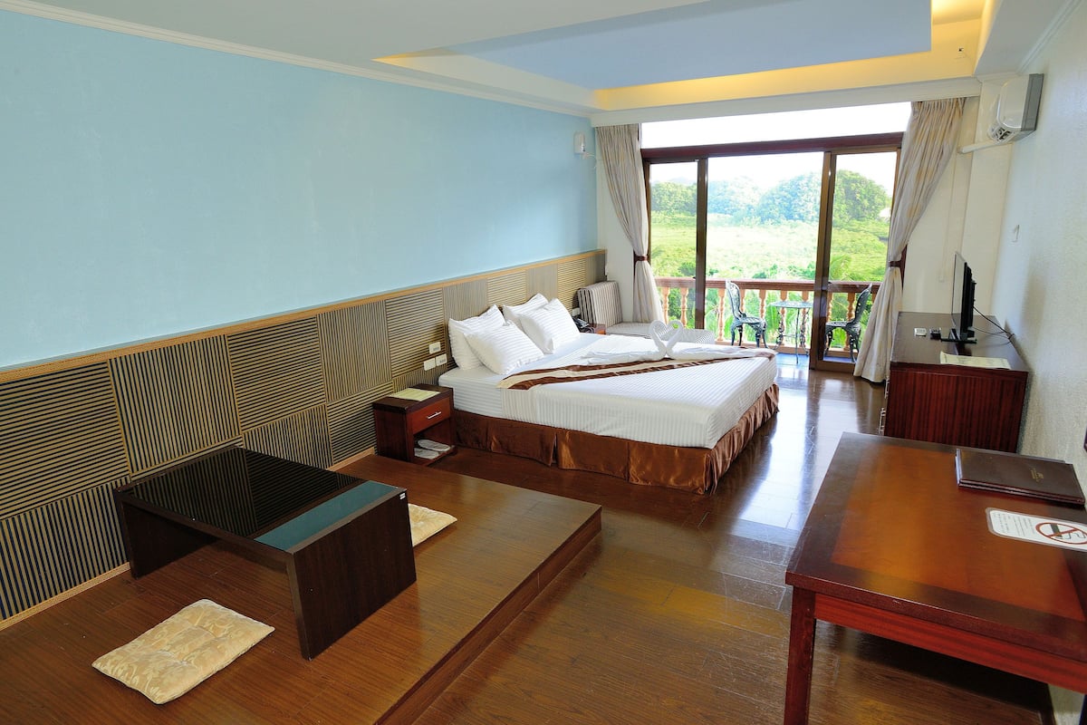 Ein Hotelzimmer mit einem großen Bett, einem Holztisch, einem kleinen Tisch und einem Balkon mit Blick auf Grünfläche.