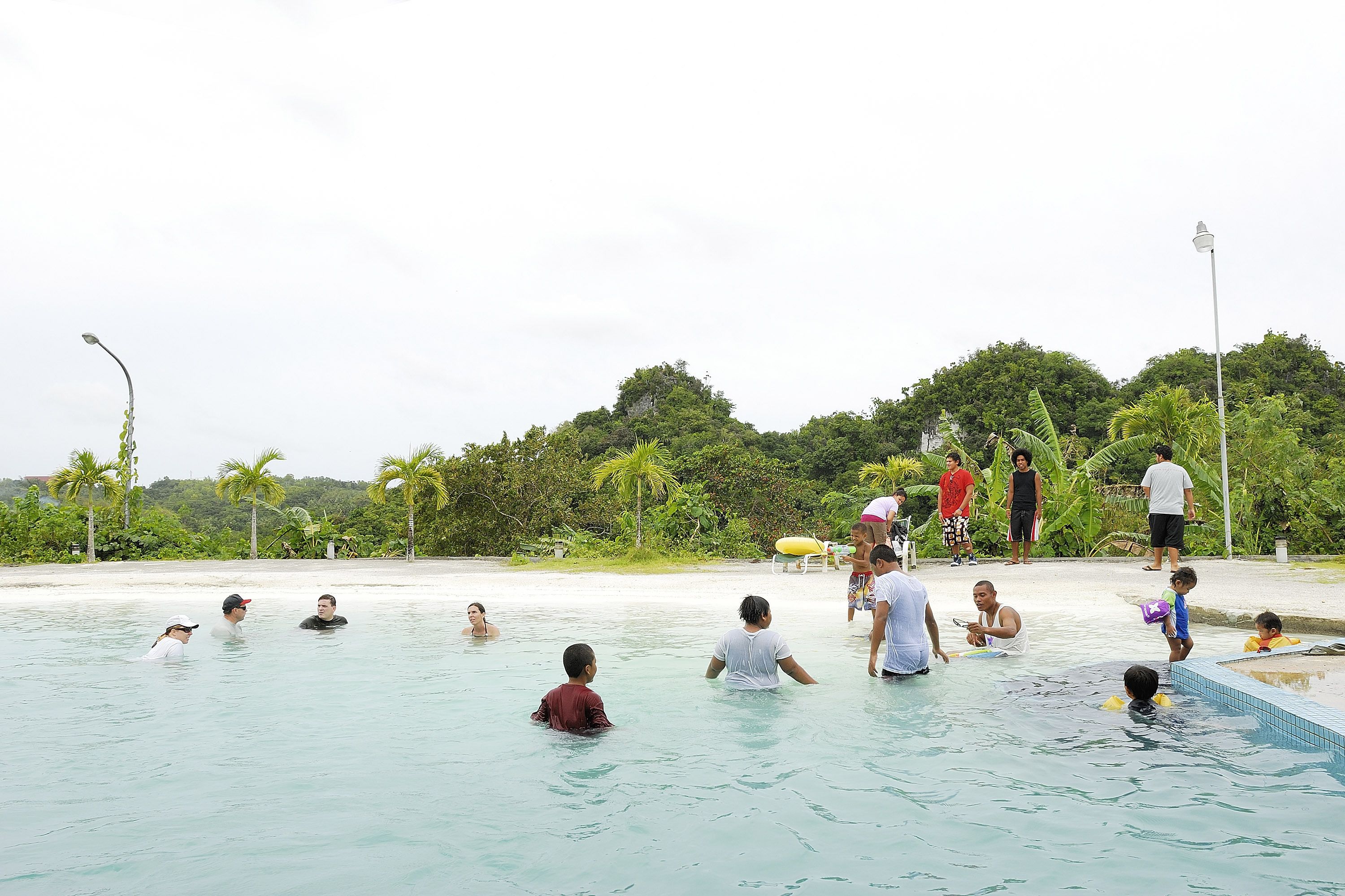 Una piscina con gente dentro y alrededor, rodeada por una playa y palmeras.