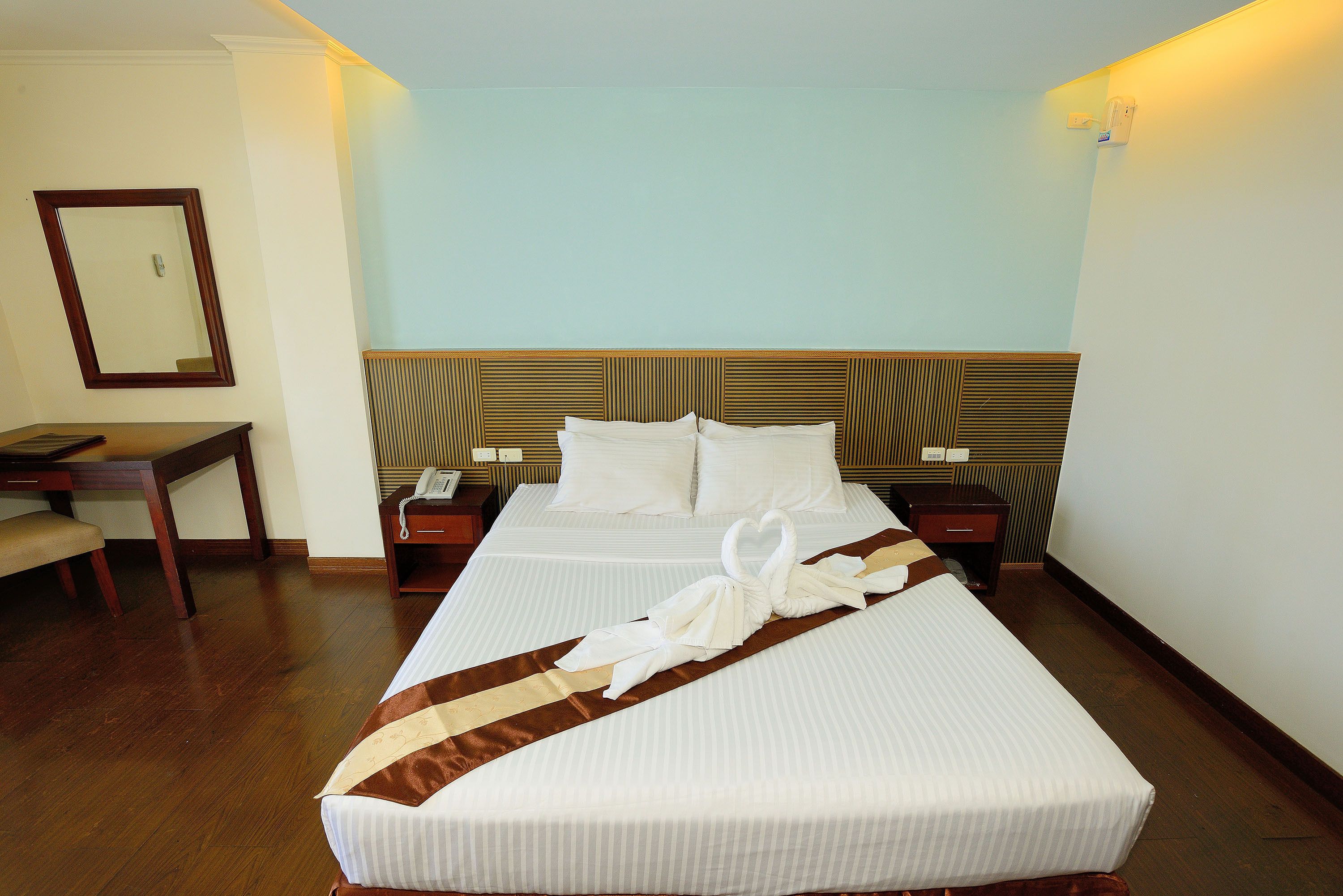Quarto de hotel com duas camas, uma cabeceira de madeira, mesa de cabeceira com abajur, espelho e um cobreleito riscado em castanho.