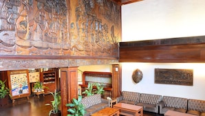 Un vestíbulo amplio con techo alto, paneles de madera y un gran mural. Hay zonas de estar con sofás y mesas, y plantas en macetas distribuidas por la sala.
