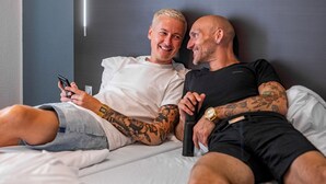 Two people lying on a bed with tattoos, one holding a phone.