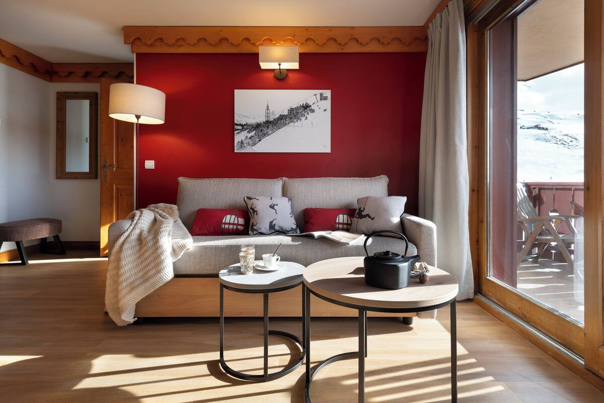 Ein gemütliches Wohnzimmer mit einer Couch, zwei Beistelltischen, einer Lampe und Blick auf eine verschneite Landschaft.