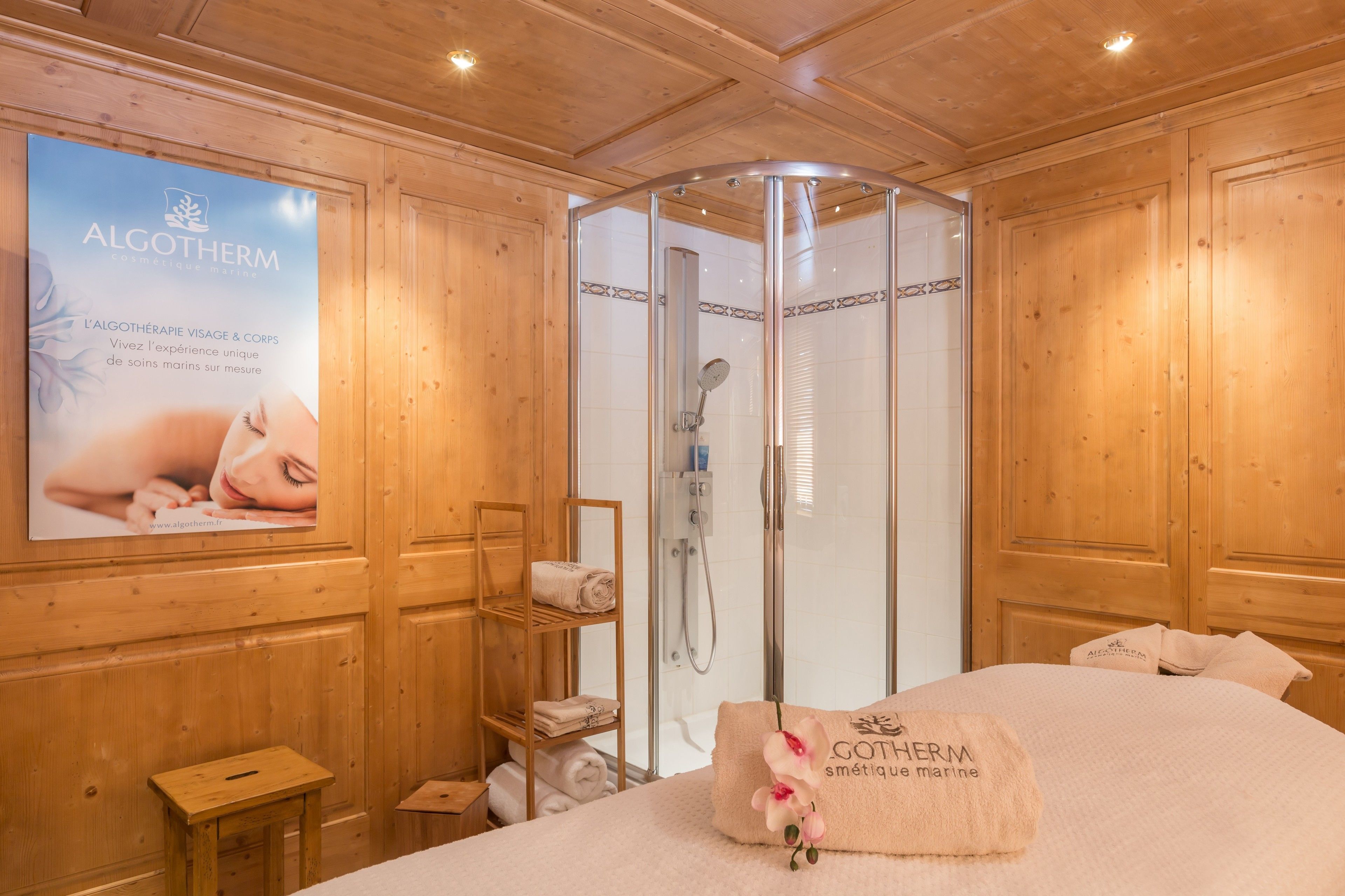 A wooden room with a glass-enclosed shower, a bed with a towel, and a poster on the wall.