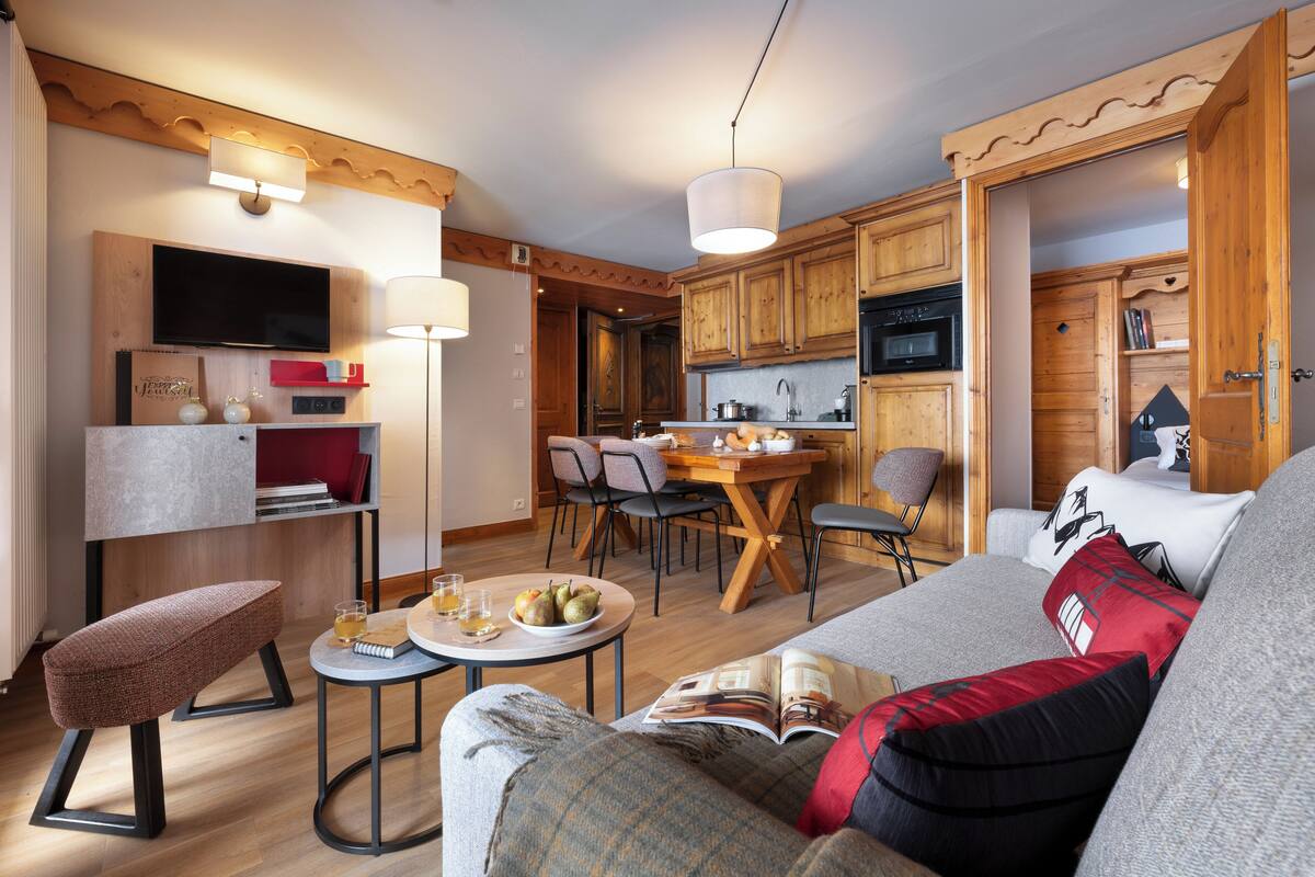 Ein modernes Wohnzimmer mit einer Couch, einem Couchtisch und einem Essbereich mit Stühlen. Im Hintergrund ist eine Küche mit Holzschränken und einem Fernseher zu sehen.