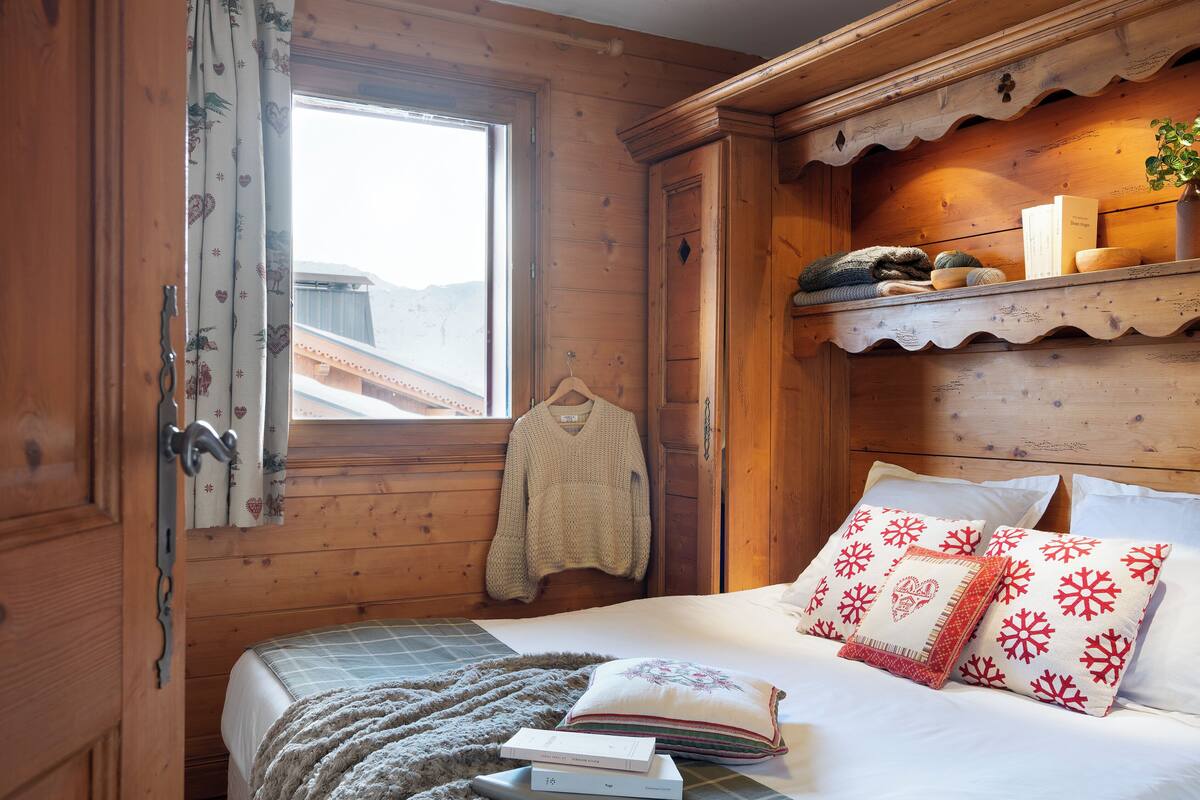 Ein gemütliches Zimmer mit Holzwänden, einem Bett mit weißer Bettwäsche und gemusterten Kissen, einem Fenster mit Vorhängen und einem Regal mit Büchern und einer Pflanze.