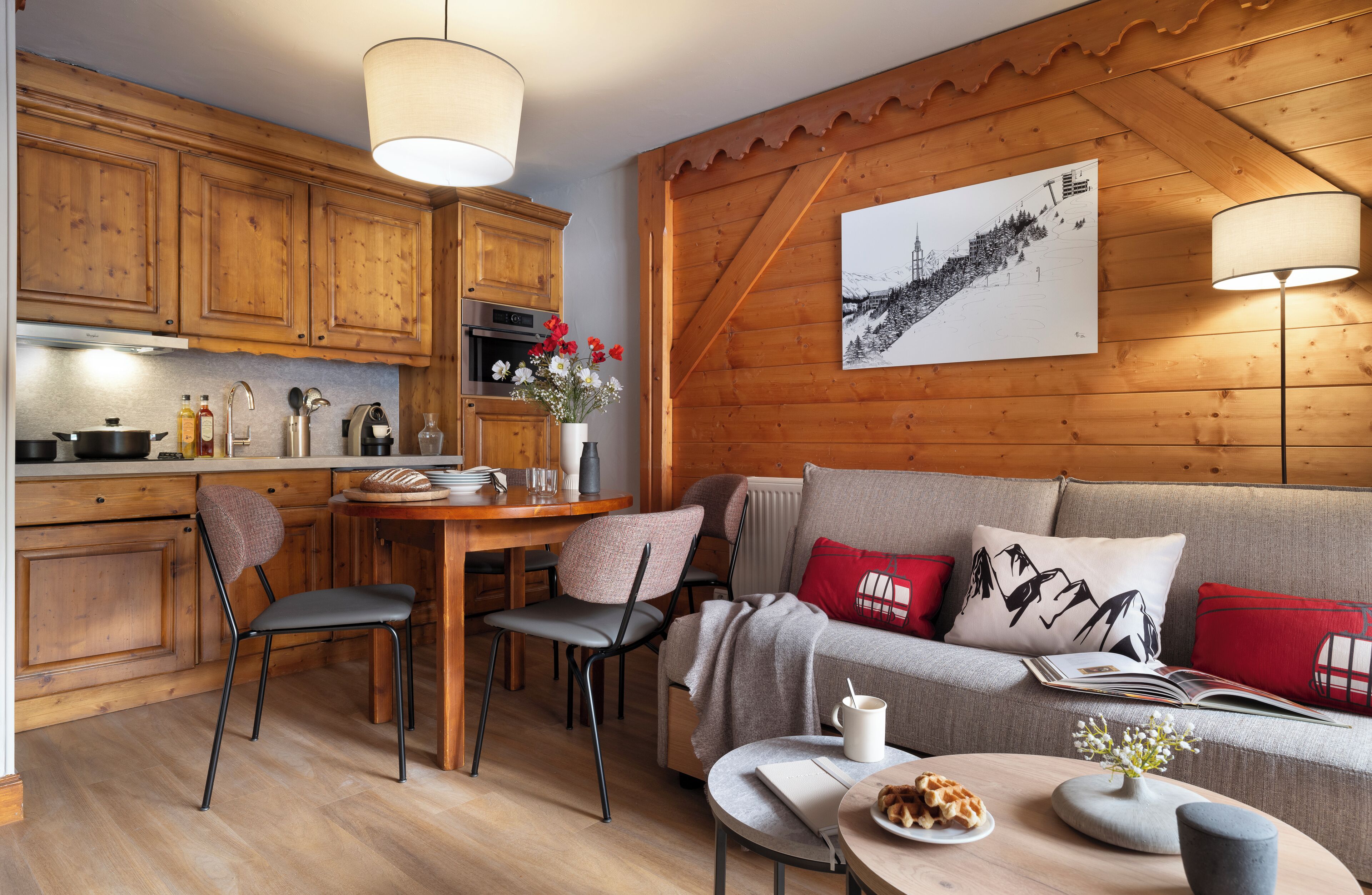 A cozy kitchen with wooden cabinets, a dining area with chairs, a table with a vase of flowers, and a sofa with red pillows.