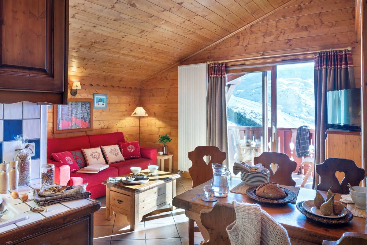 Ein gemütliches Holzhaus-Interieur mit einem roten Sofa, einem Esstisch und Blick auf verschneite Berge durch große Fenster.