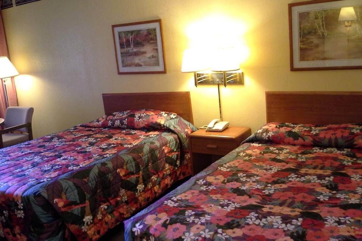 A hotel room with two beds, a nightstand with a lamp, a phone, and a chair.