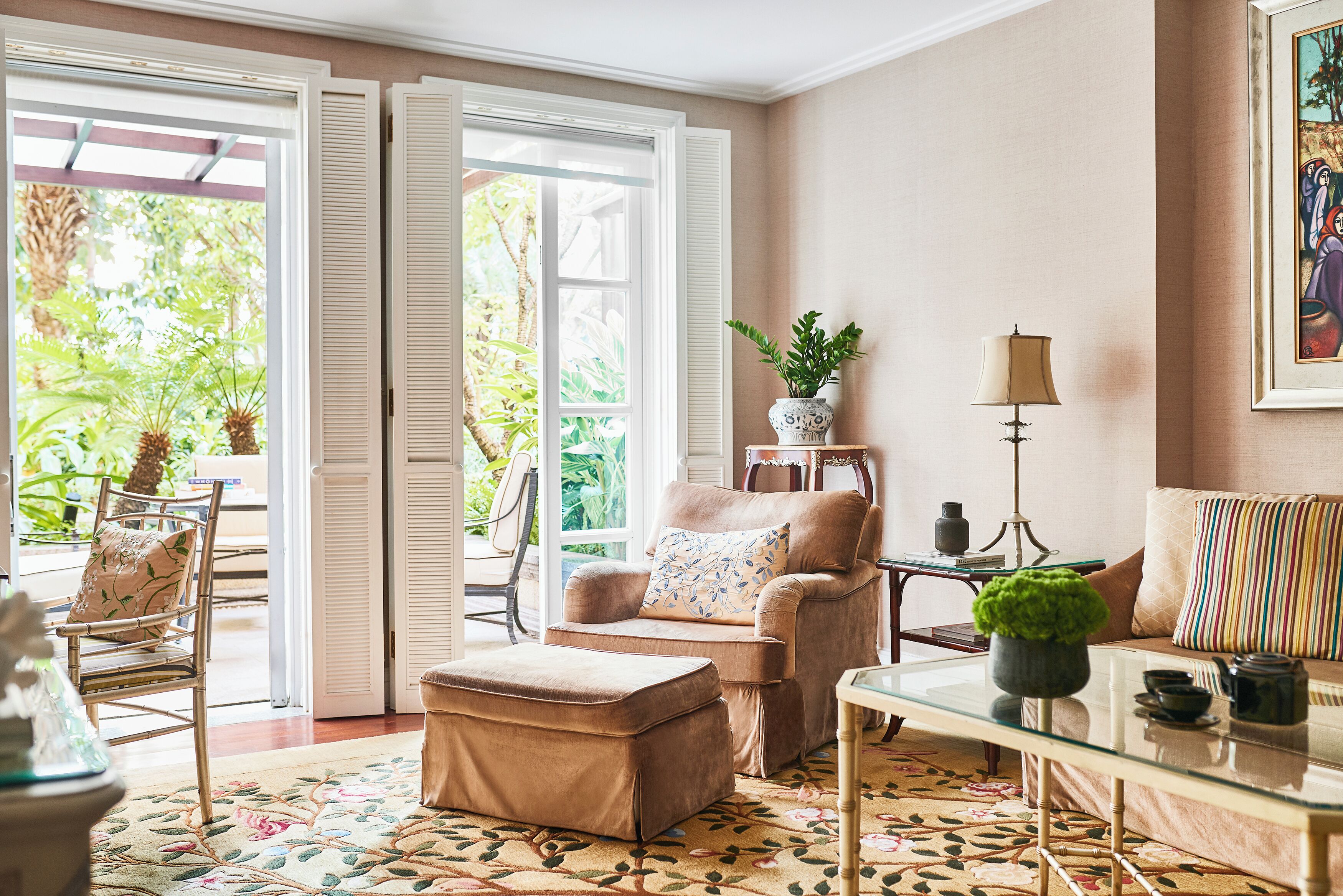 A room with a sofa, armchair, ottoman, and a table with a lamp. There is a window with shutters, a plant on the table, and a painting on the wall.
