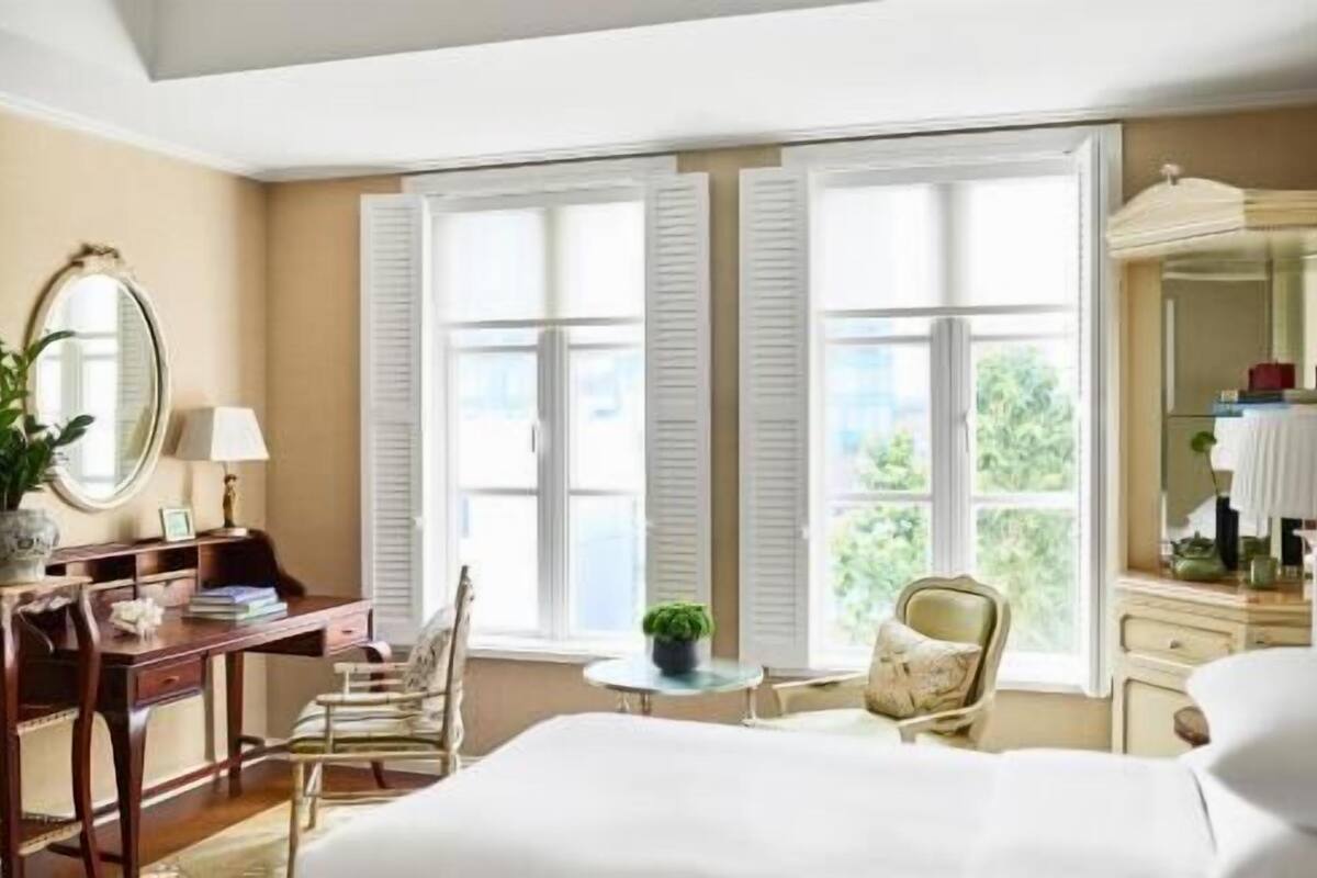 Une chambre d’hôtel comprenant un lit, un bureau, une chaise, une table ronde et une grande fenêtre avec des volets.