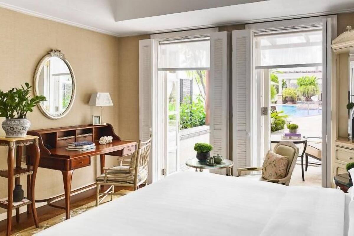 Une chambre d’hôtel comprenant un lit, un bureau avec une chaise, une petite table avec une chaise, un miroir et une vue sur une piscine et la verdure environnante.