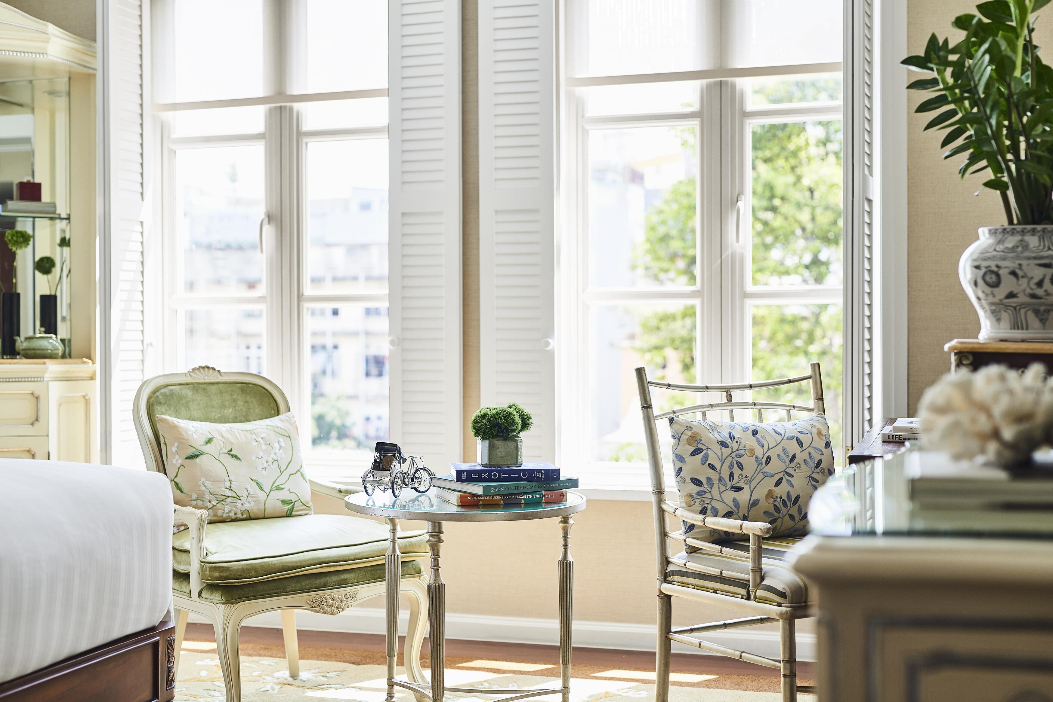 A well-lit room with a bed, a chair, a small table with books and a plant, and a window with blinds.
