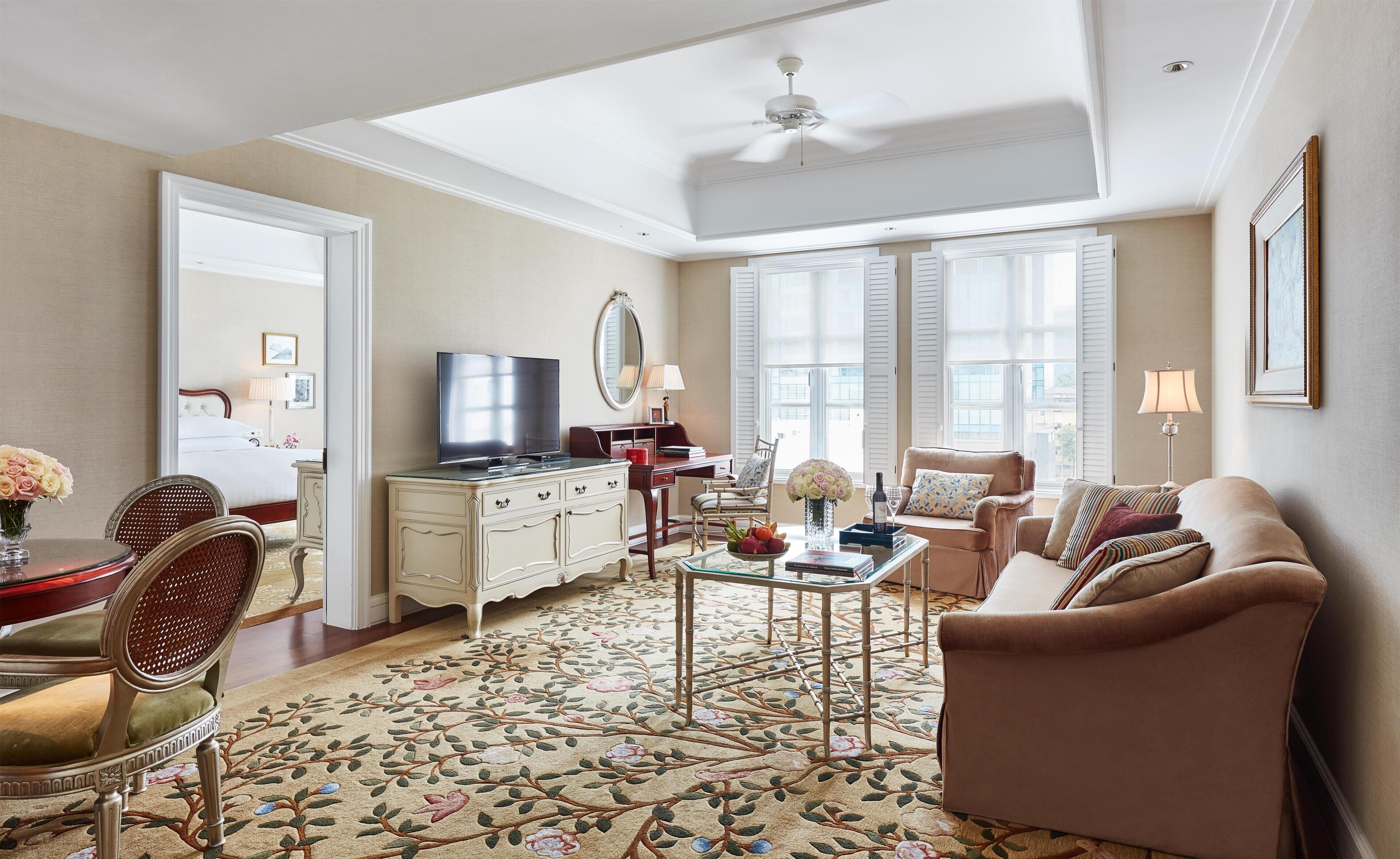 A spacious living room with a large window, a ceiling fan, a flat-screen TV, a sofa, a coffee table, and a decorative rug.