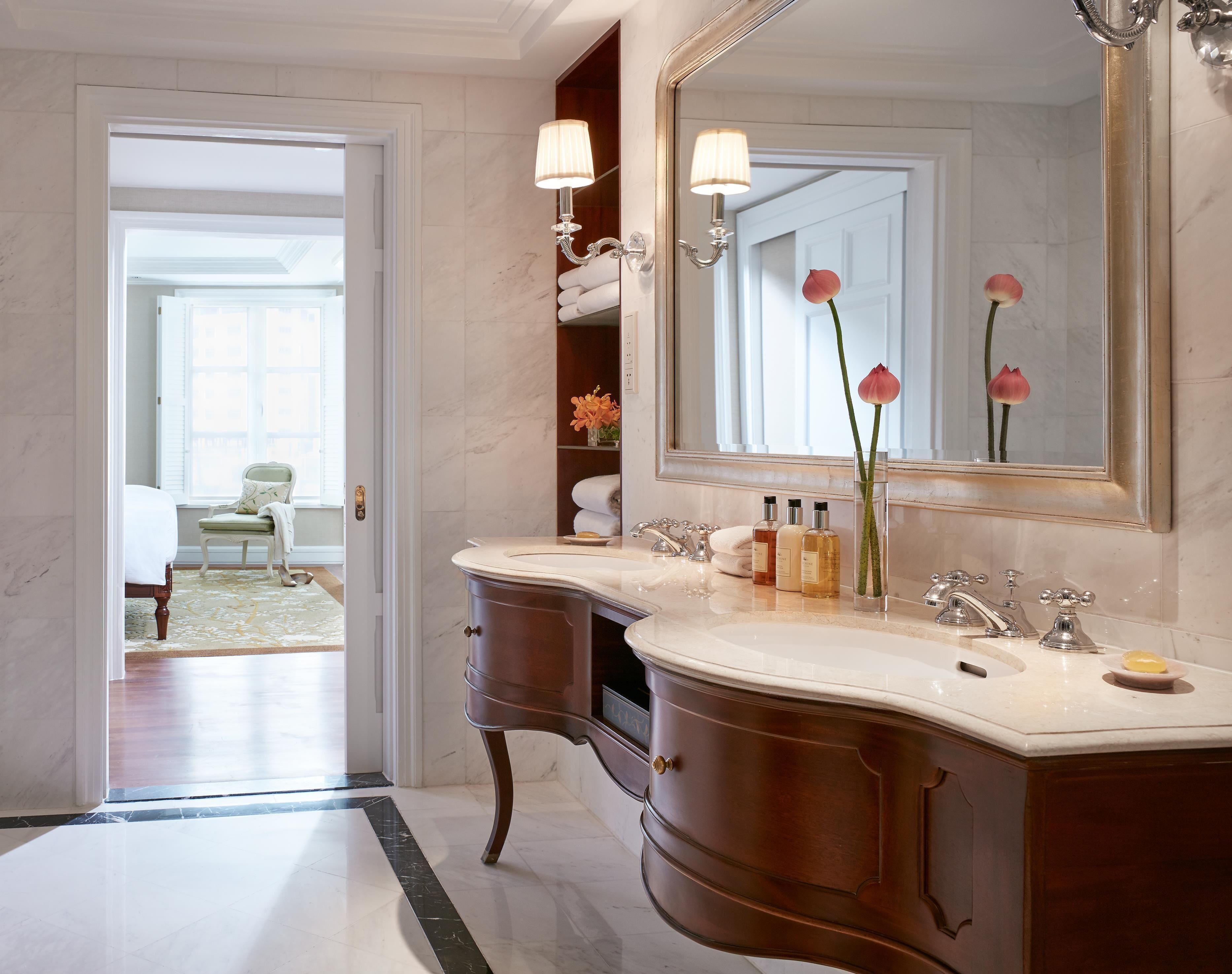 A bathroom with a double vanity, a large mirror, and a view into a bedroom.