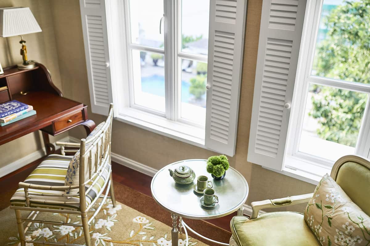 Une pièce avec un bureau en bois, une chaise, une table ronde avec une théière et des tasses, et une vue sur la verdure par une fenêtre à volets.