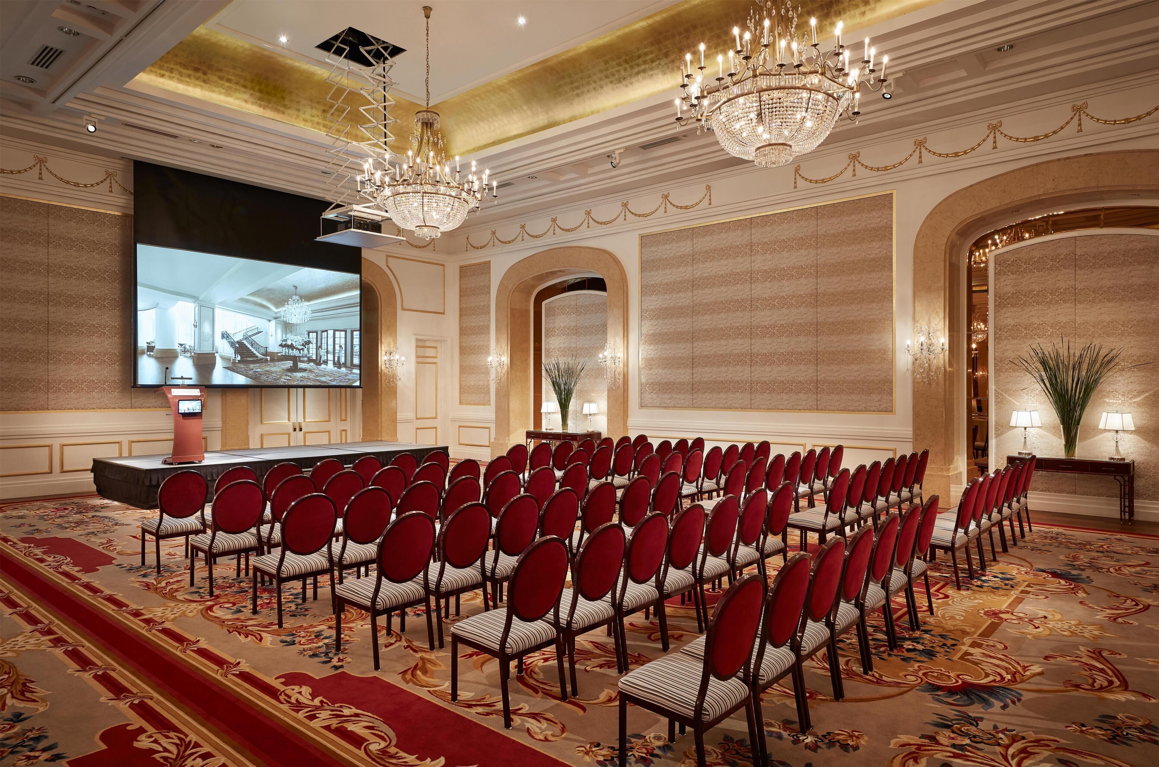 A conference room with rows of chairs, a large screen, and chandeliers.