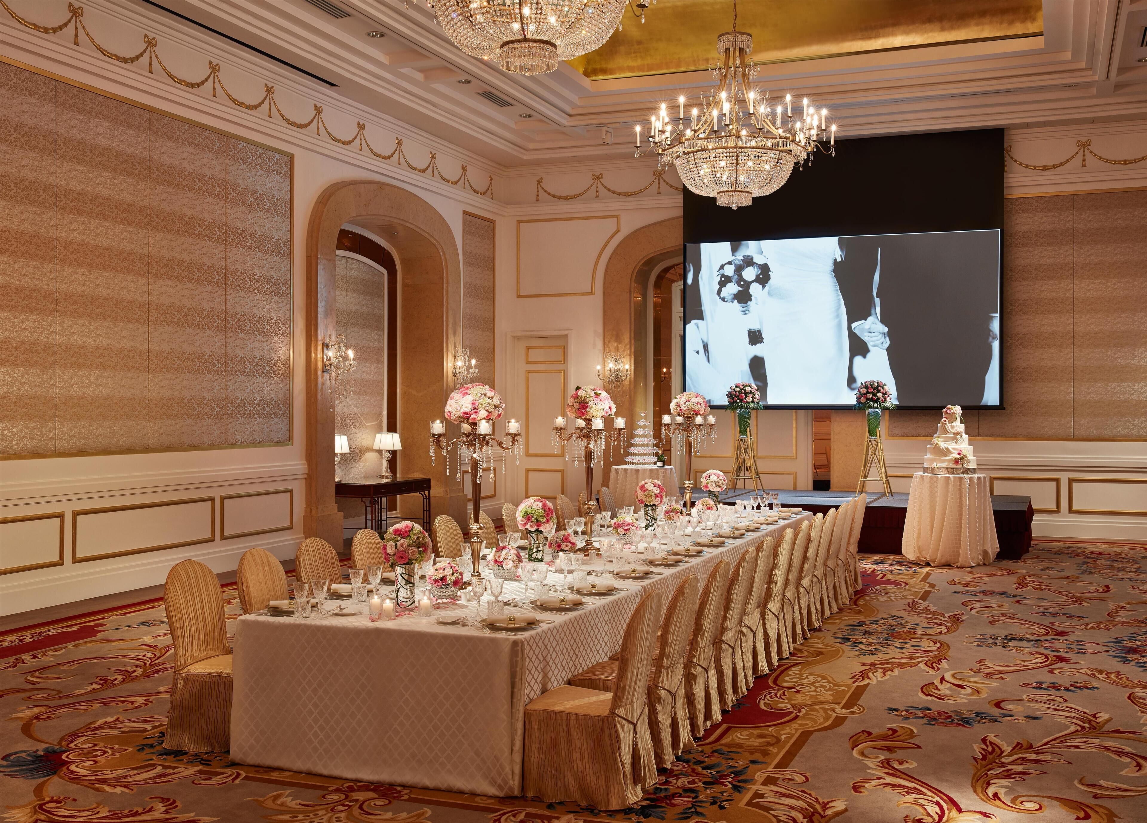 A banquet hall with a long table set for a formal event, chandeliers, and a large screen displaying a wedding.