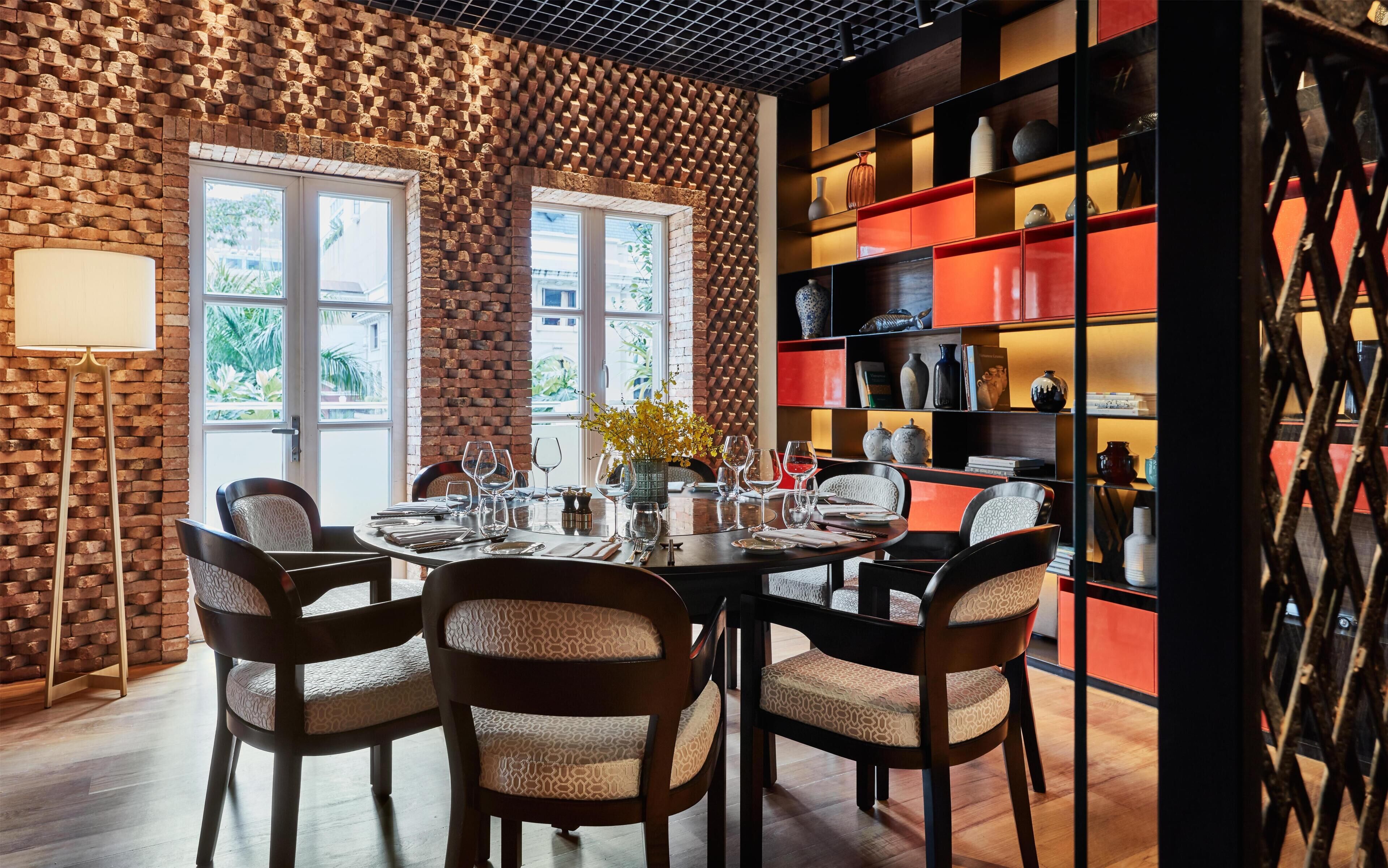 Uma sala de jantar com parede de tijolos, mesa redonda posta para seis pessoas e uma estante com diversos objetos decorativos.