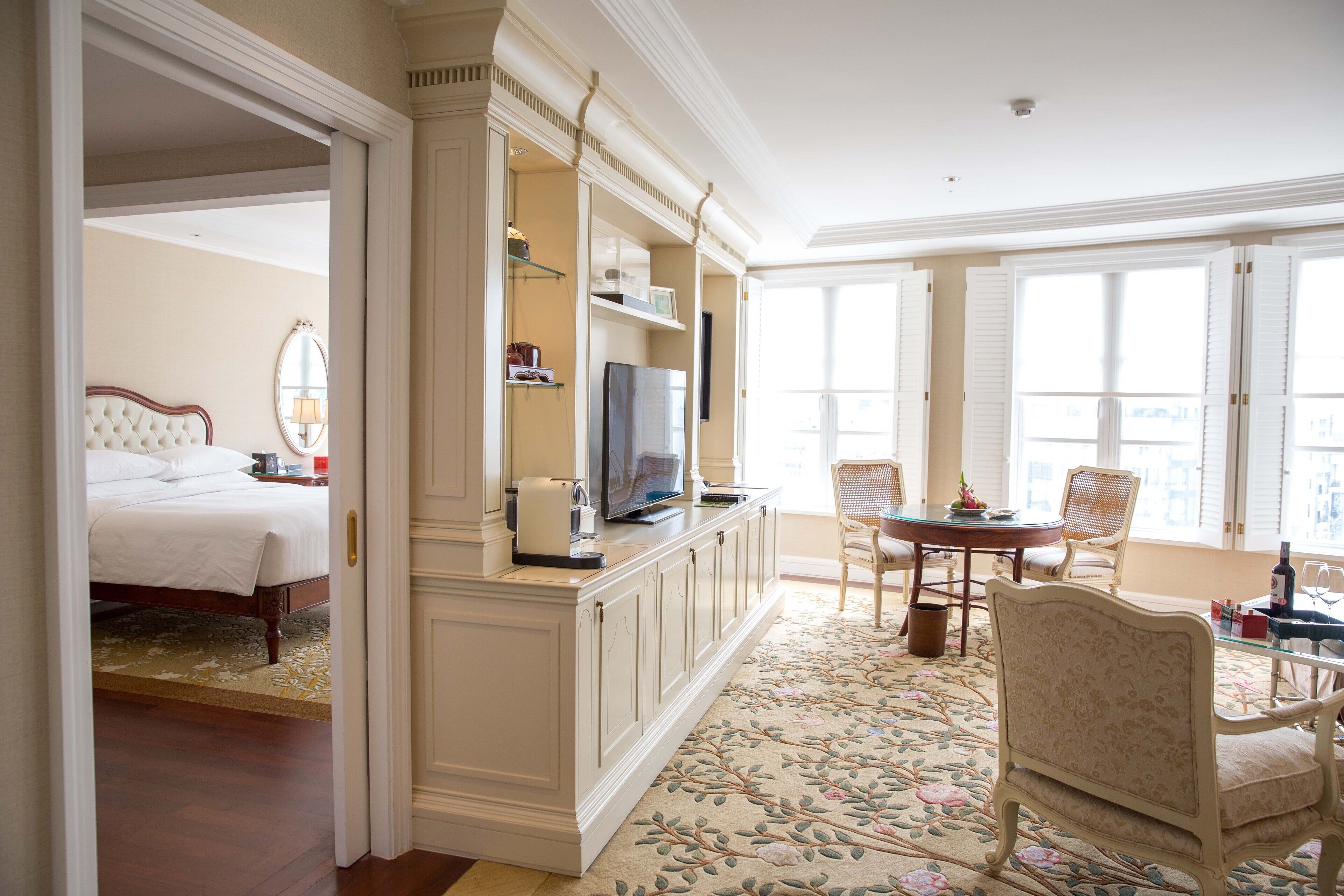 A hotel room with a bed, a TV on a stand, a dining table with chairs, and a large window with shutters.