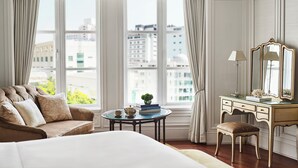 A hotel room with a large bed, a sitting area with a sofa and armchair, a small round table with a lamp and books, and a dressing table with a mirror.