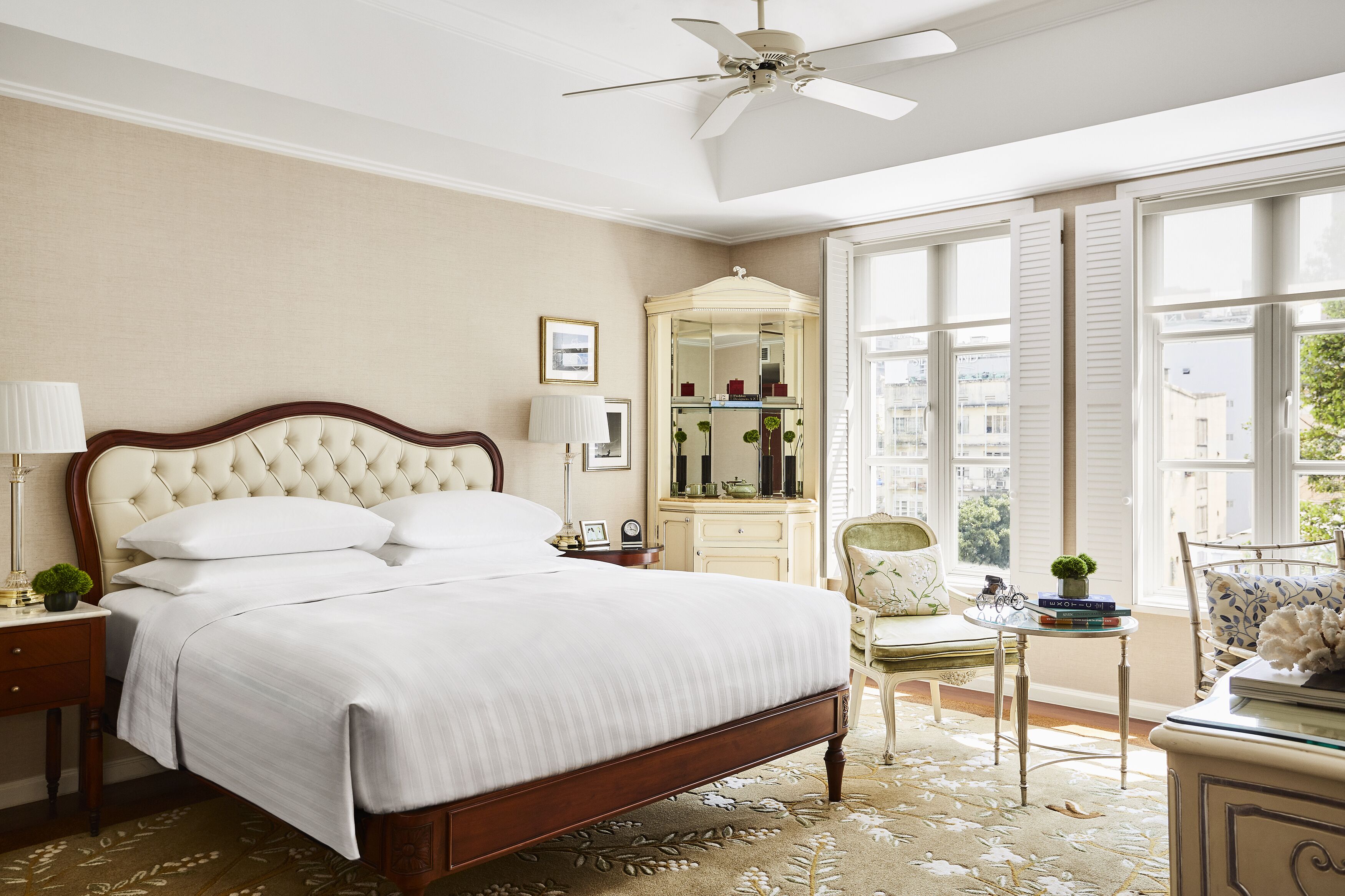 A neatly arranged bedroom with a large bed, a bedside table with a lamp, a chair, a small table with books and a plant, and a mirror.