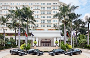 A multi-story hotel with a grand entrance, palm trees, and four black luxury cars parked in front.