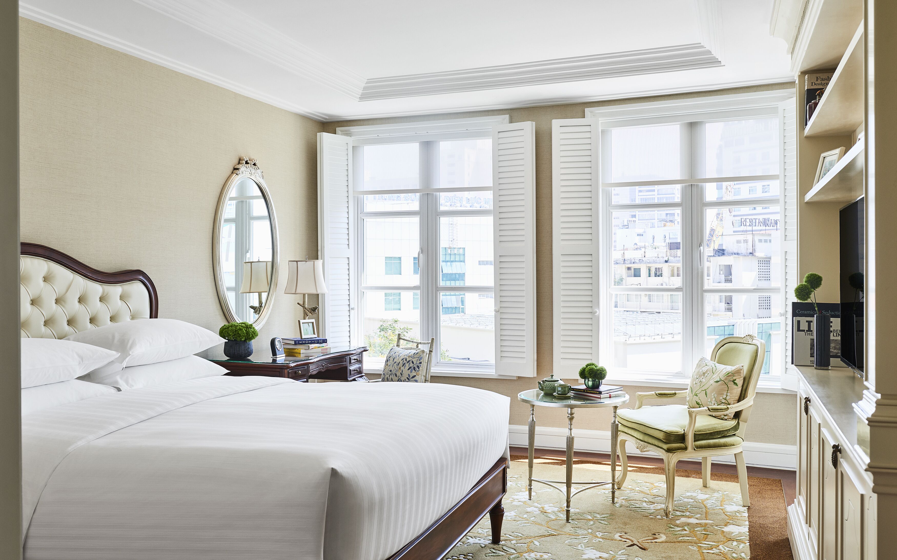 A hotel room with a large bed, a sitting area with a chair and table, a mirror, and a view of the city through large windows.