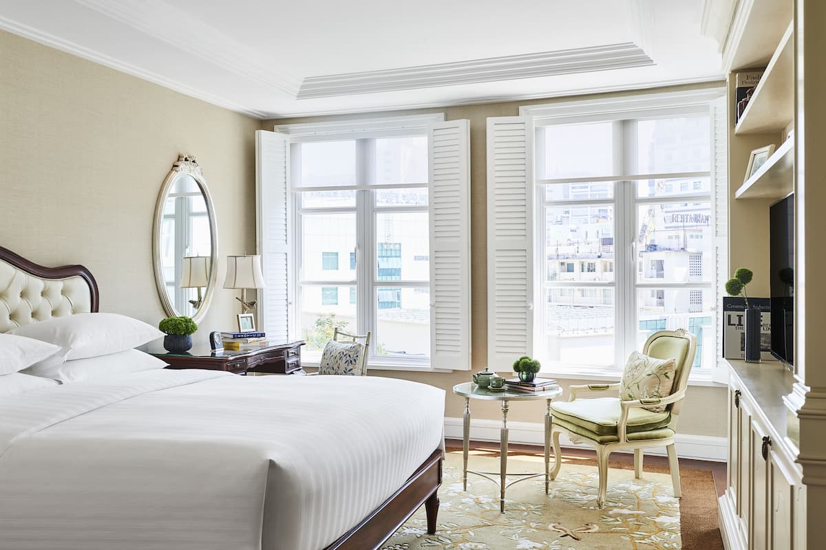 Une chambre d’hôtel dotée d’un grand lit, d’un coin salon avec un fauteuil et une table, d’un miroir et offrant une vue sur la ville grâce à de grandes fenêtres.