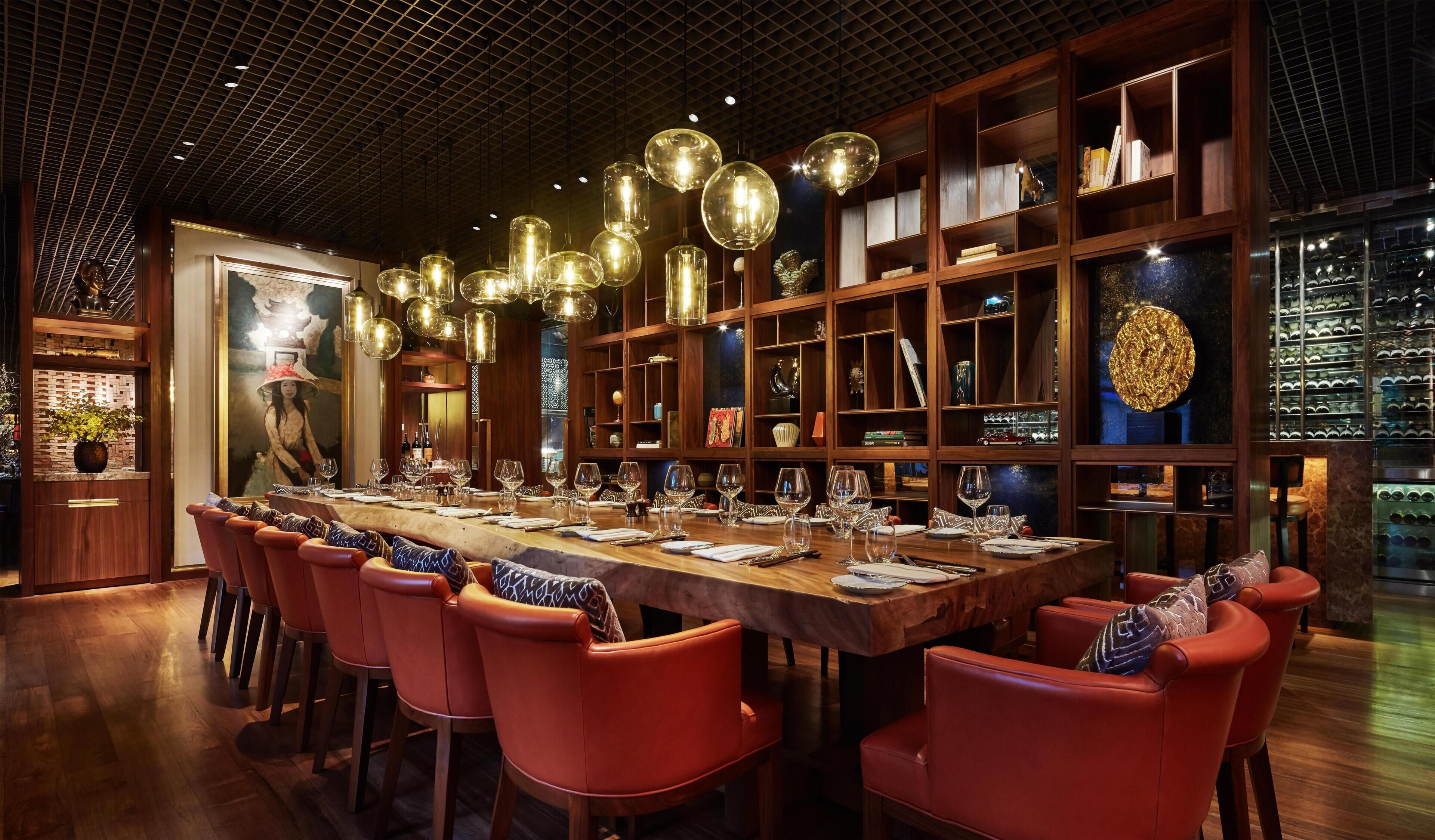 A restaurant with a long wooden table set for dining, red upholstered chairs, and a wall of shelves with various items.