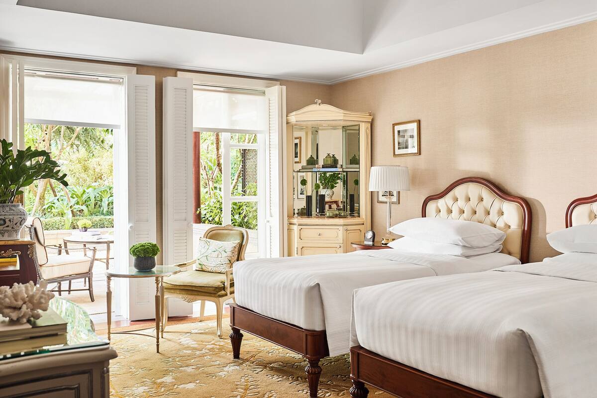 Une chambre d’hôtel avec deux lits, une coiffeuse avec un miroir, une petite table avec une chaise, et une vue sur la verdure.