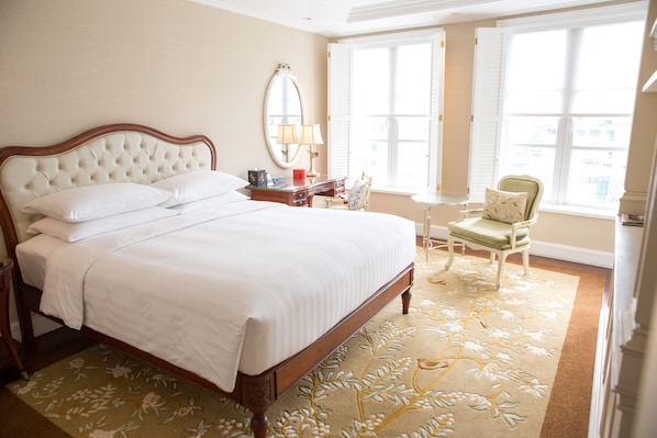 Una cama bien hecha con cabecera de madera, una mesita de noche con lámpara, una silla y un ventanal con contraventanas.