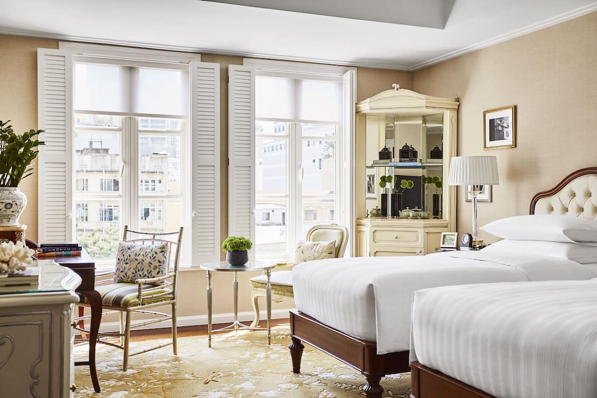 Une chambre d’hôtel avec deux lits, une grande fenêtre avec des volets, une coiffeuse avec un miroir et une petite table avec une plante.