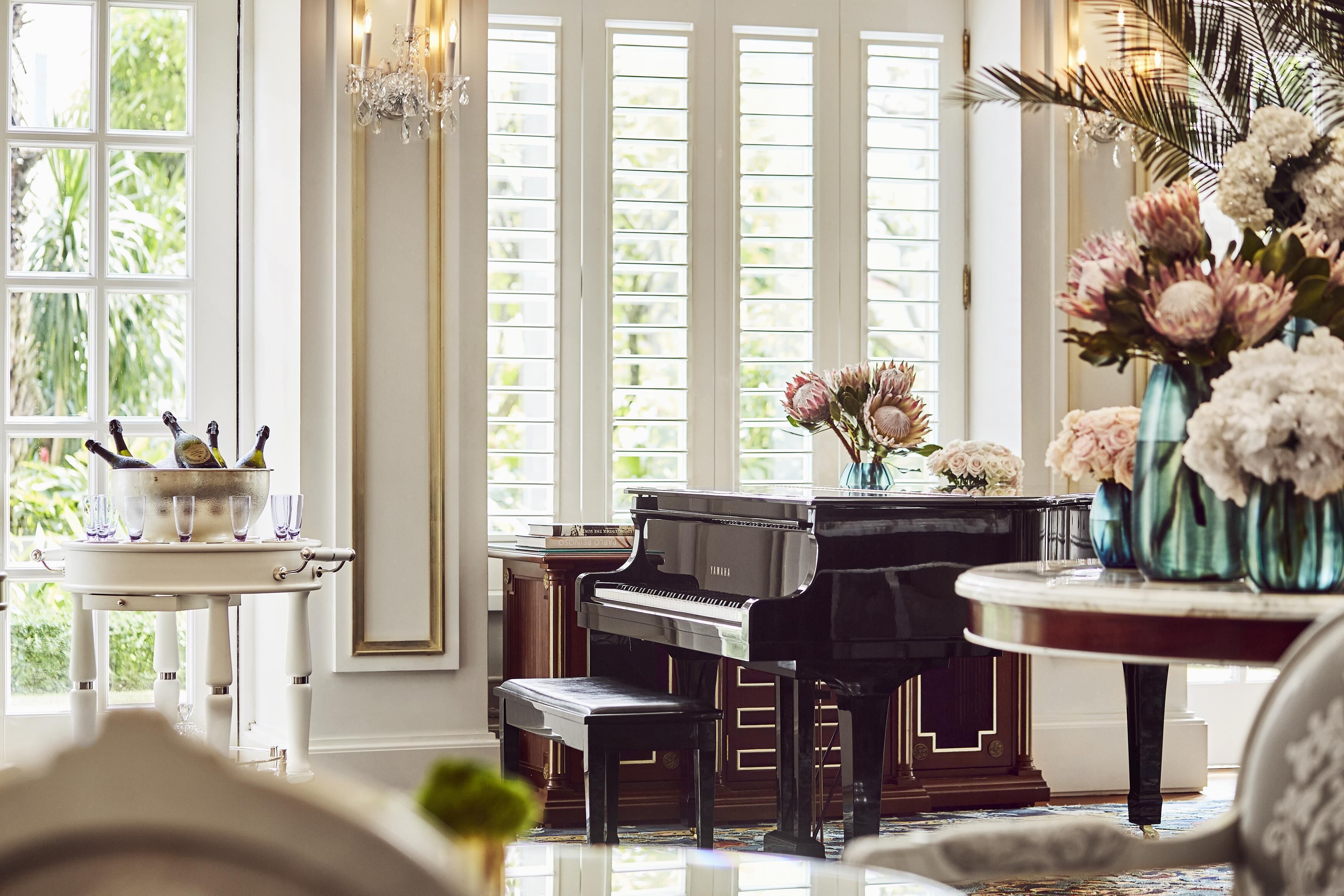 Un piano de cola en una habitación luminosa con ventanas amplias, una araña y un florero con flores.