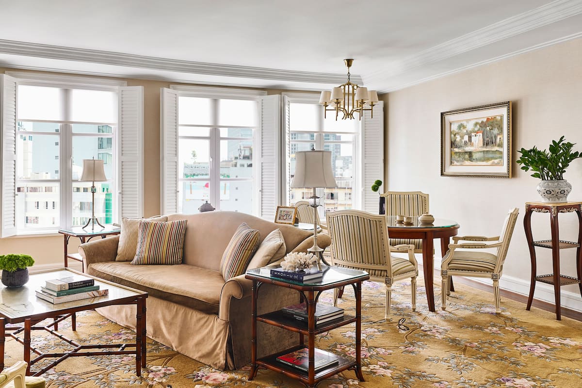 Un salon spacieux avec un canapé beige, une table basse et un tapis à motifs floraux. De grandes fenêtres sont dotées de volets blancs, et une suspension orne le plafond. Un tableau encadré est accroché au mur.