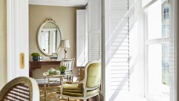 Une pièce bien éclairée, dotée d’un grand miroir, d’un fauteuil recouvert d’un tissu vert, d’une petite table avec une lampe et d’une vue sur l’extérieur grâce à une fenêtre ornée de volets blancs.