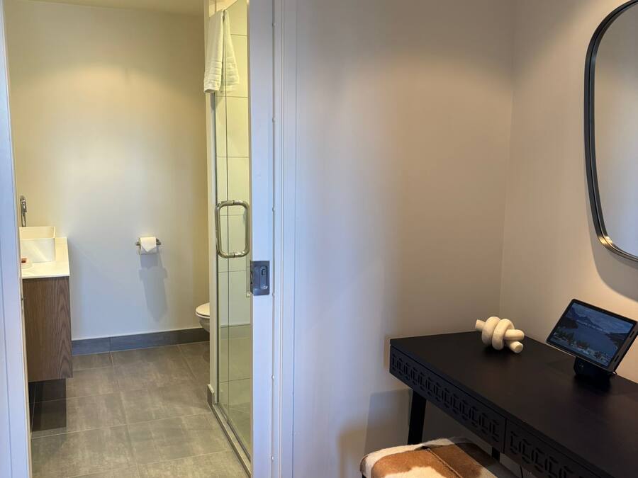 A hotel room with a desk, a bench, a mirror, and a view of the bathroom.