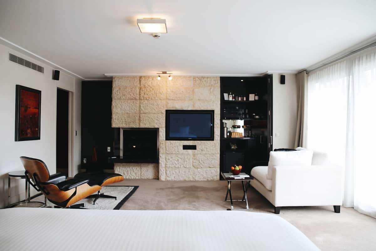Una habitación de hotel moderna con una cama grande, una zona de estar con sillones, un televisor de pantalla plana montado en una pared de piedra y un minibar.