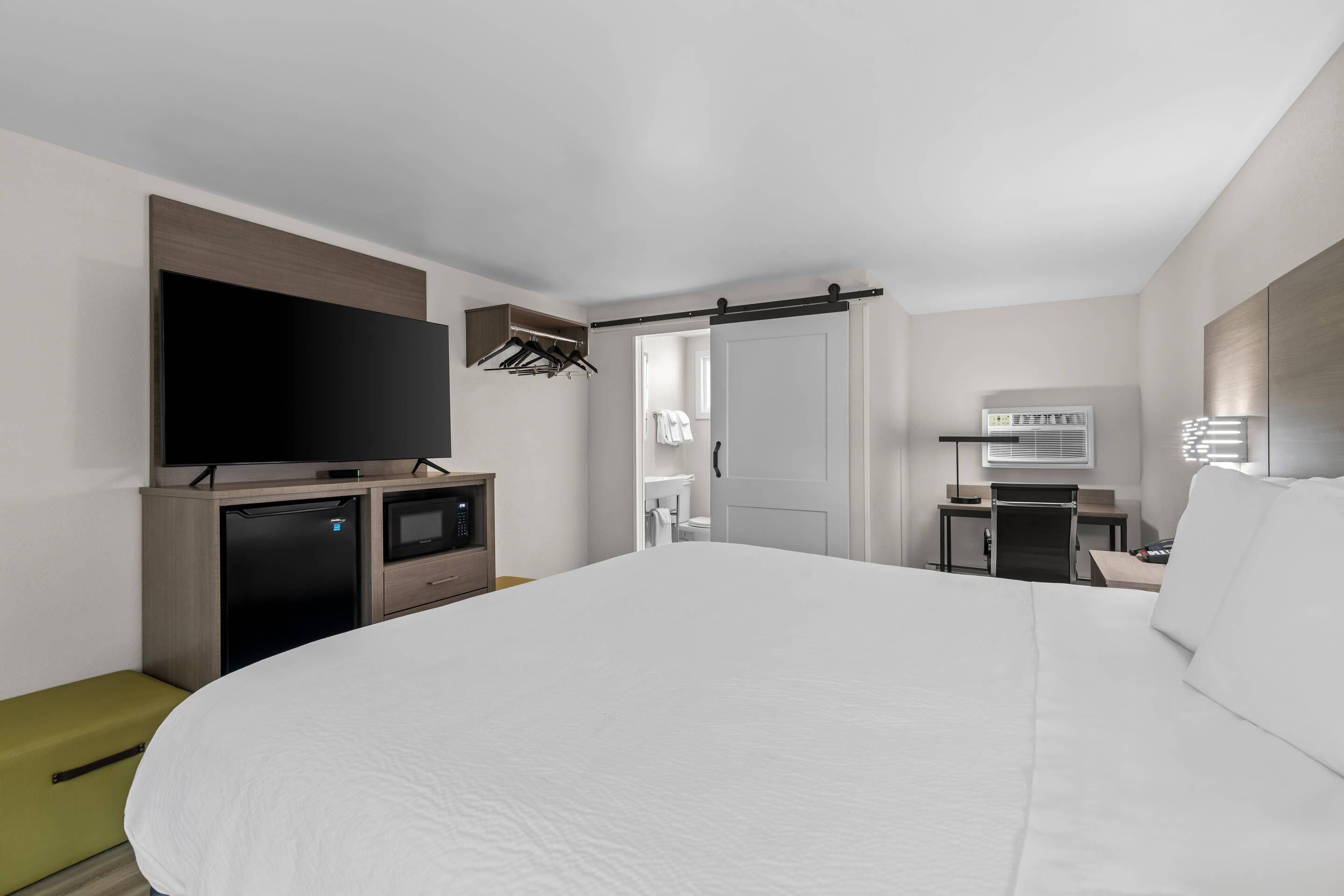 Habitación de hotel con una cama grande, televisor de pantalla plana, escritorio con silla, equipo de aire acondicionado y puerta corredera que da a un baño.