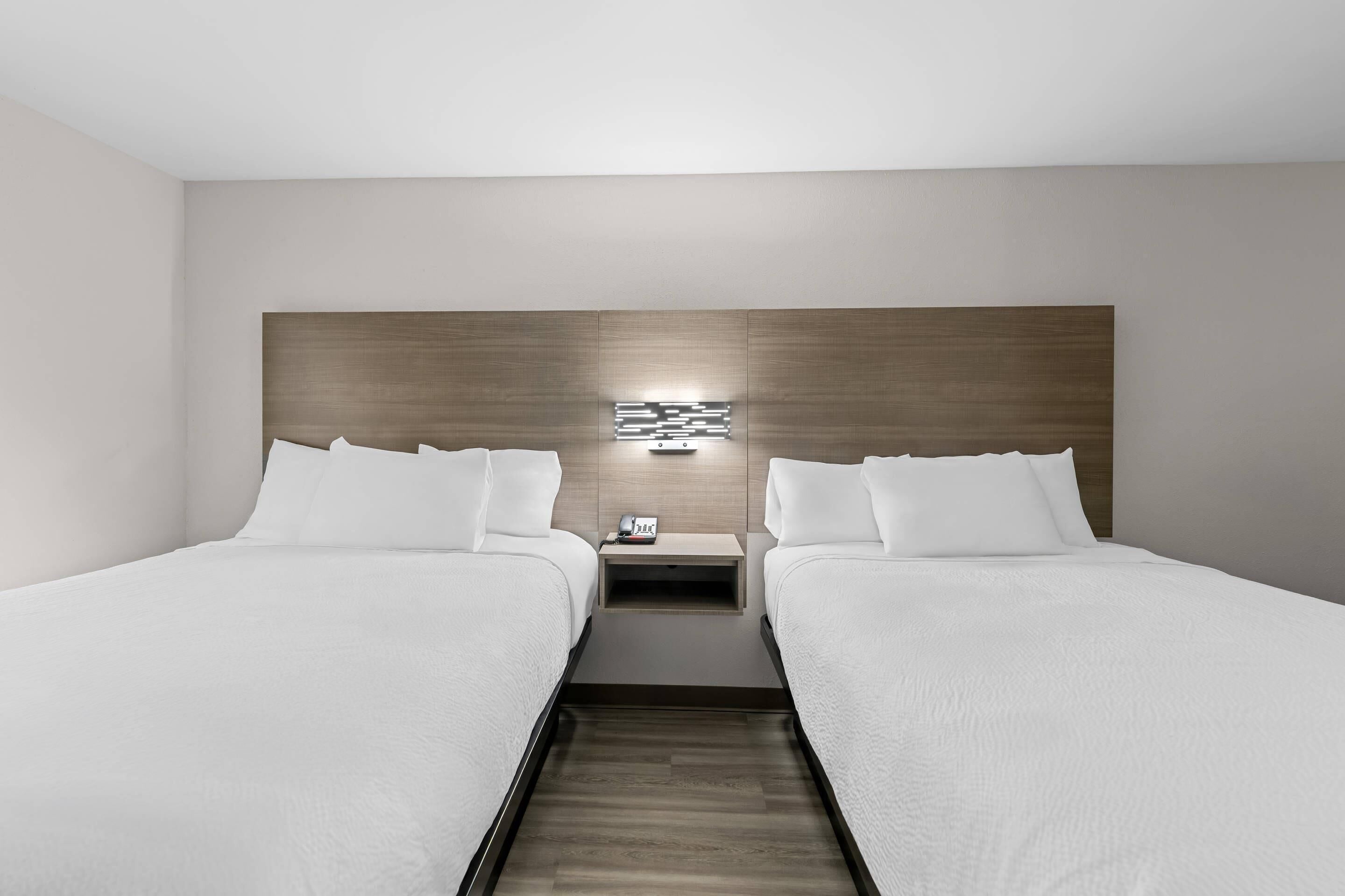 A modern hotel room with two beds, a wooden headboard, a nightstand with a phone, and a wall-mounted light.