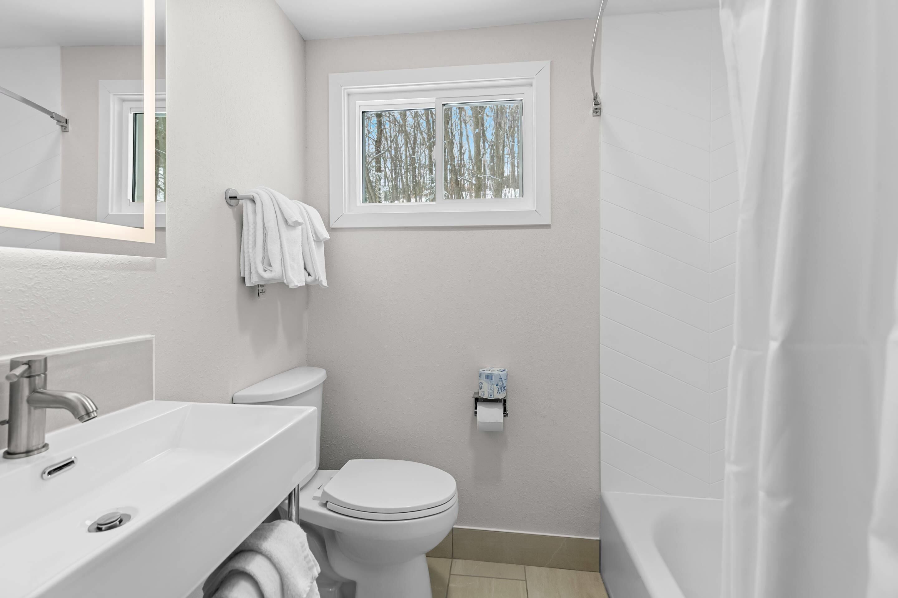 A bathroom with a white sink, toilet, and bathtub. There is a window with a view of trees and a towel hanging on the wall.