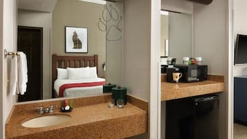 A hotel room with a bed, a bathroom with a sink and mirror, and a kitchenette with a microwave and coffee maker.